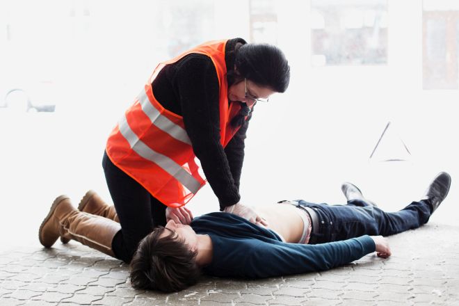 Mann liegt am Boden. Ersthelferin kniet daneben. Pullover des Mannes ist hoch gezogen. Auf dem nackten Oberkörper wird eine Herzdruckmassage durchgeführt. 