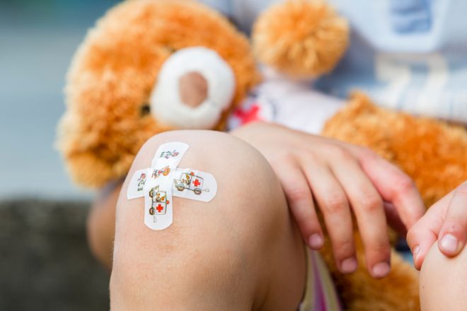 Kind sitzend, auf einem Knie sind zwei Pflaster in Kreuz-Form aufgebracht. In der Hand hält das Kind einen Plüsch-Teddy mit DRK-Shirt.