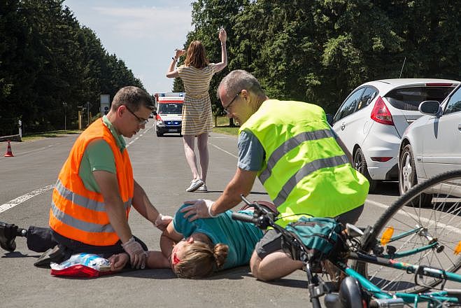 Fahrradunfall auf einer Straße. Fahrradfahrer liegt am Boden. Neben ihm knien zwei Ersthelfende, die jeweils eine Warnweste tragen. Im Vordergrund sieht man ein Fahrrad liegen. Eine weist den Rettungswagen ein, der auf die Unfallstelle zufährt.